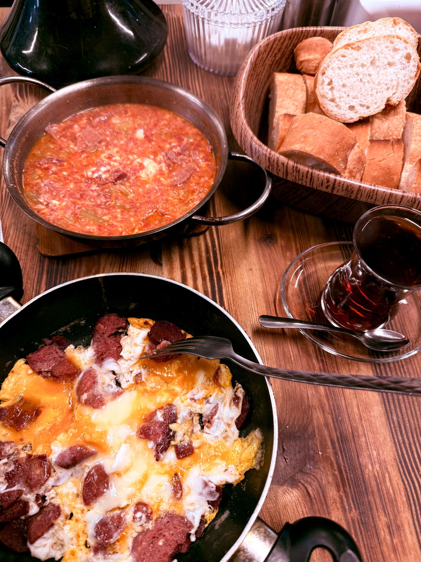 A pan of tomato-and-egg menemen and a pan of sucuklu yumurta with cured sausage, a basket of bread and a glass of black tea on a wooden table