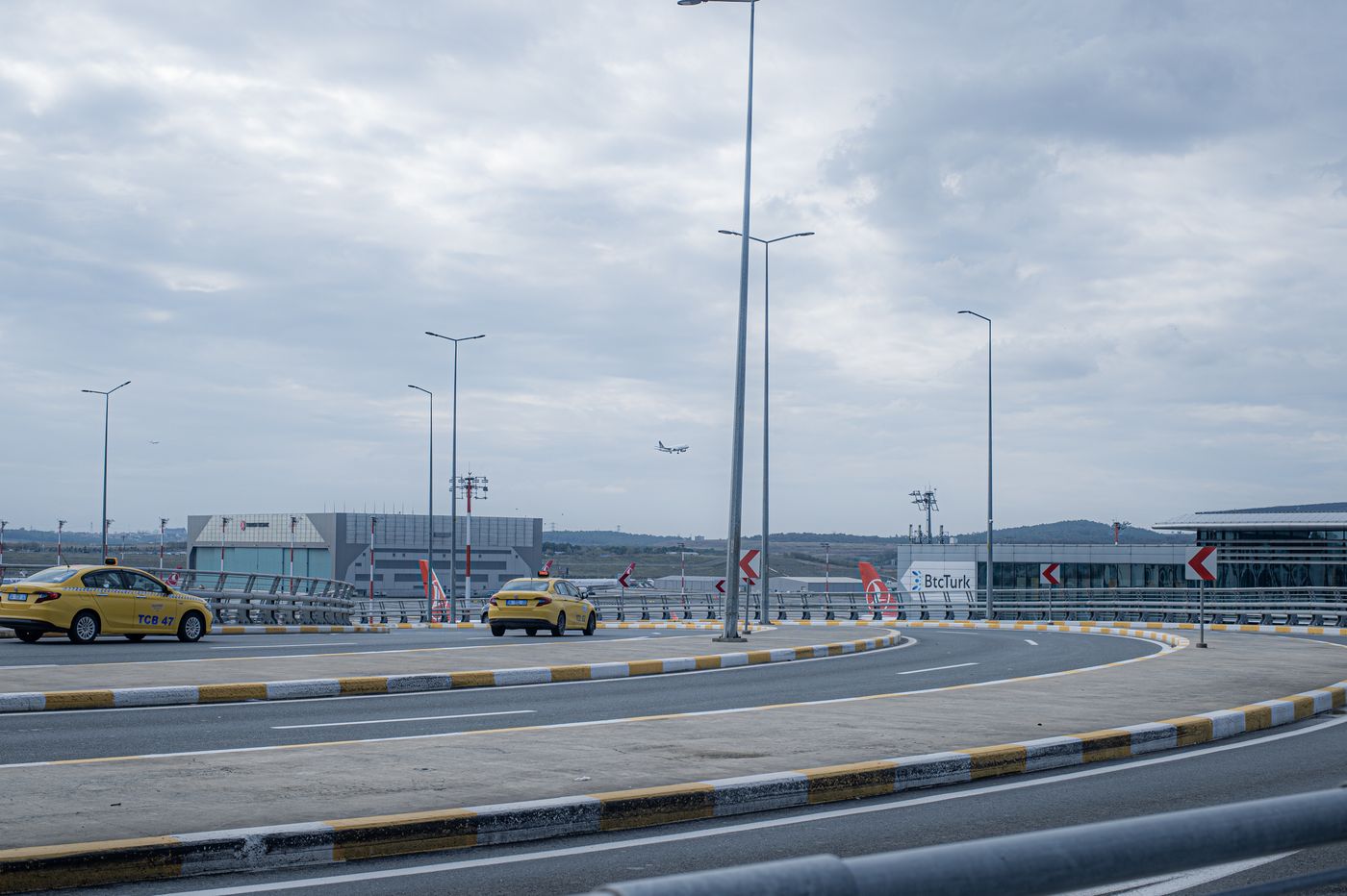 Airport access road curving past terminal buildings with a yellow Istanbul taxi and a plane on approach overhead