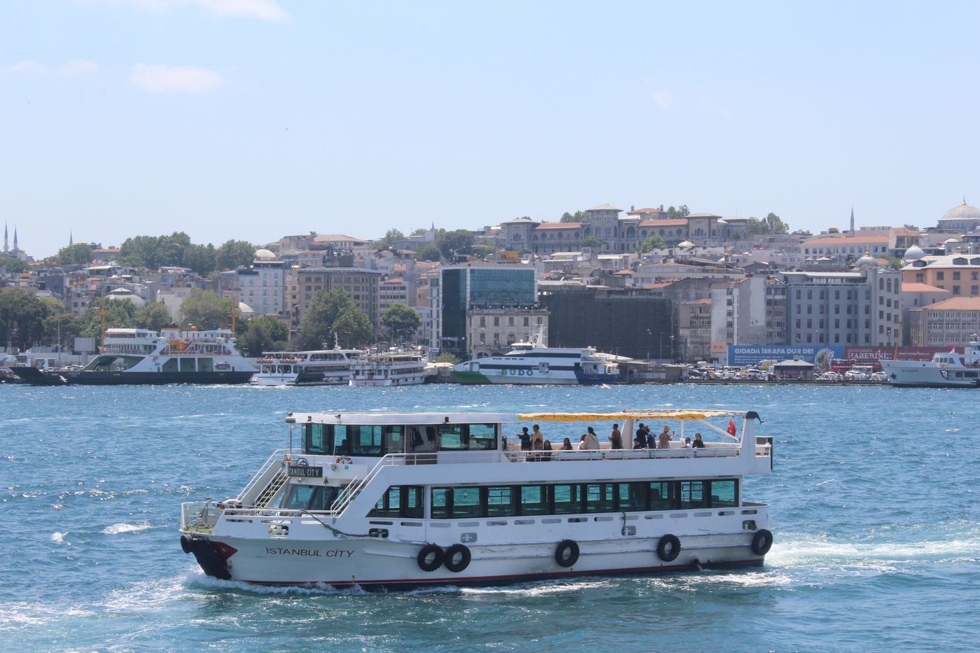 A passenger boat crossing the water in Istanbul with the historic Galata-side skyline and ferry-busy waterfront behind it