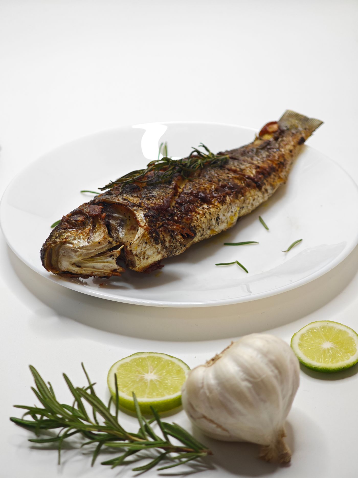 A whole grilled fish on a white plate with rosemary, lime and garlic, the dish you order at the end of a meyhane meal